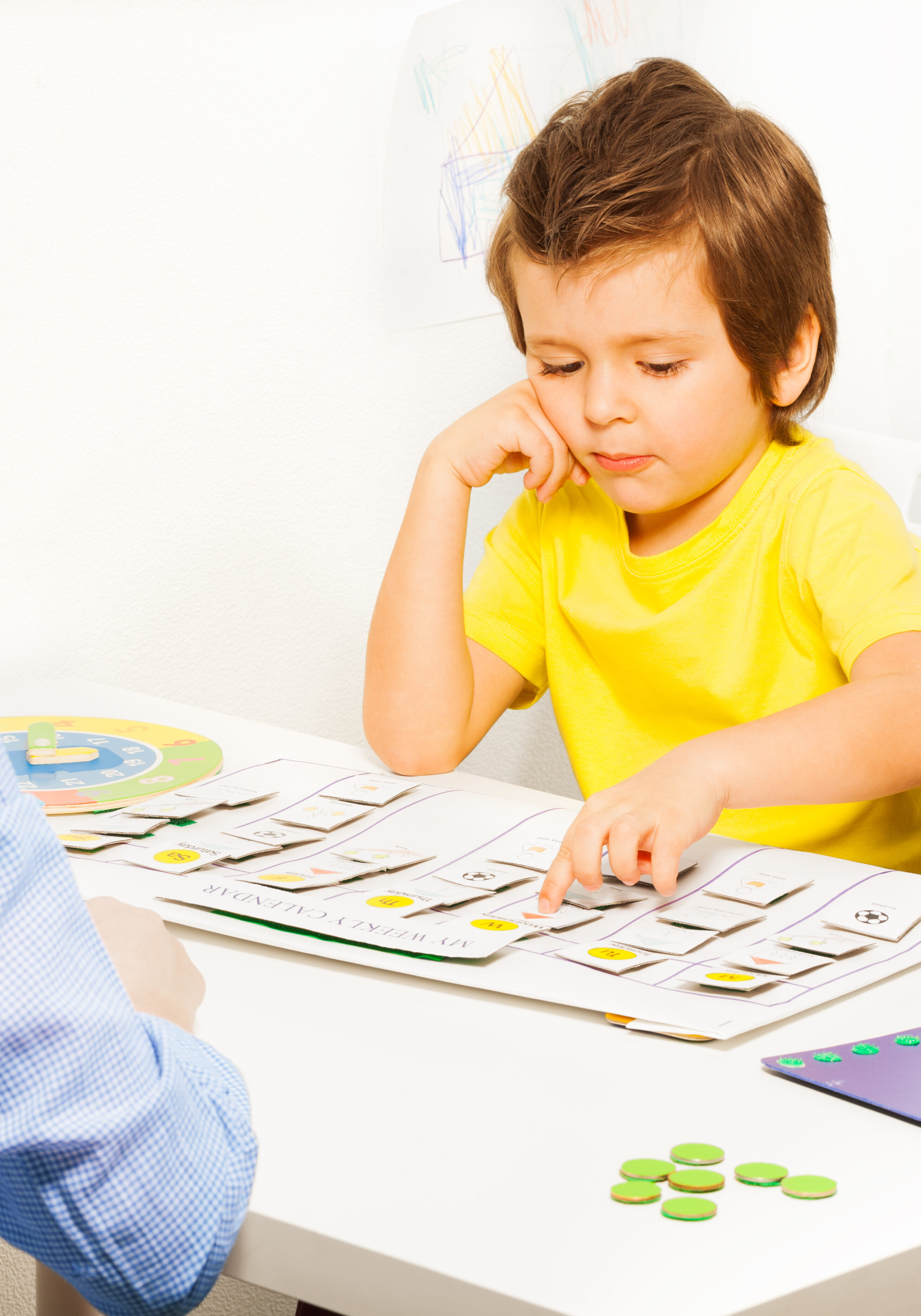 Boy pointing at colorful day activities cards on calendar during developing game with his parent sitting opposite at the table in the room